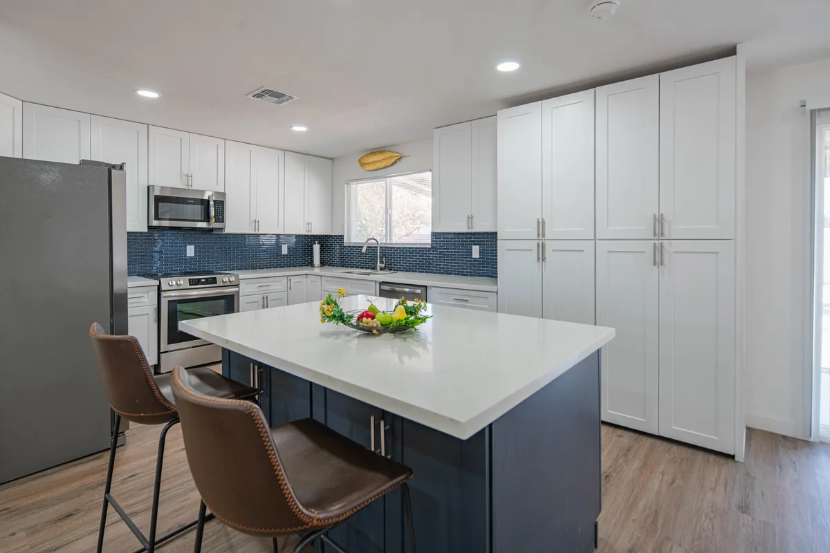 A spacious kitchen featuring a central island countertop with bar stools, a striking blue tile backsplash, and modern kitchen appliances. The setup invites both cooking and casual gatherings, with ample cabinetry visible for storage.