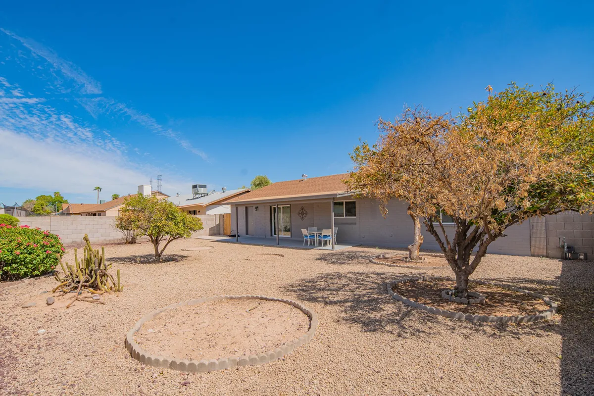 A peaceful exterior shot capturing the house with gravel landscaping, mature trees, and a welcoming patio seating area. There is ample space for outdoor activities such as family gatherings, relaxing, or letting pets roam. The image conveys a sense of openness and connection to the natural environment.