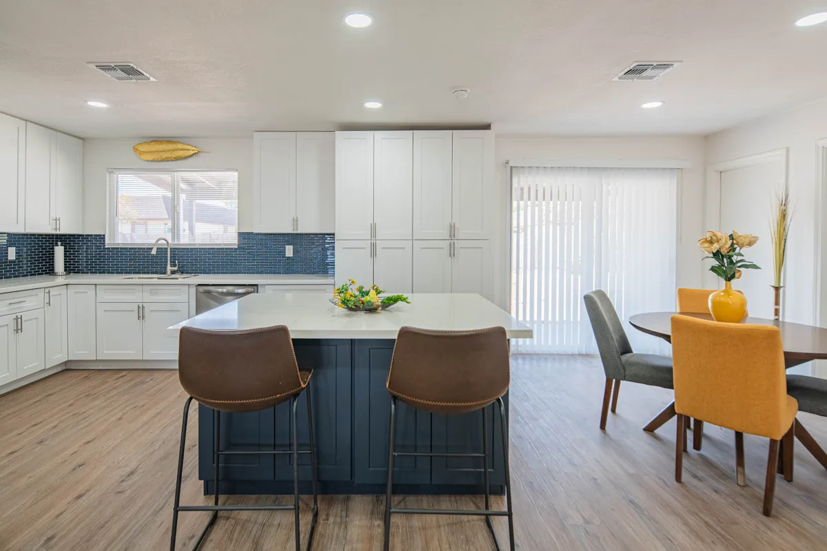 An open kitchen layout with a spacious island, integrated dining area, blue backsplash, and a beautiful flower arrangement as a centerpiece. The scene is bright and cheerful, highlighting both the kitchen workspace and the connected dining space.