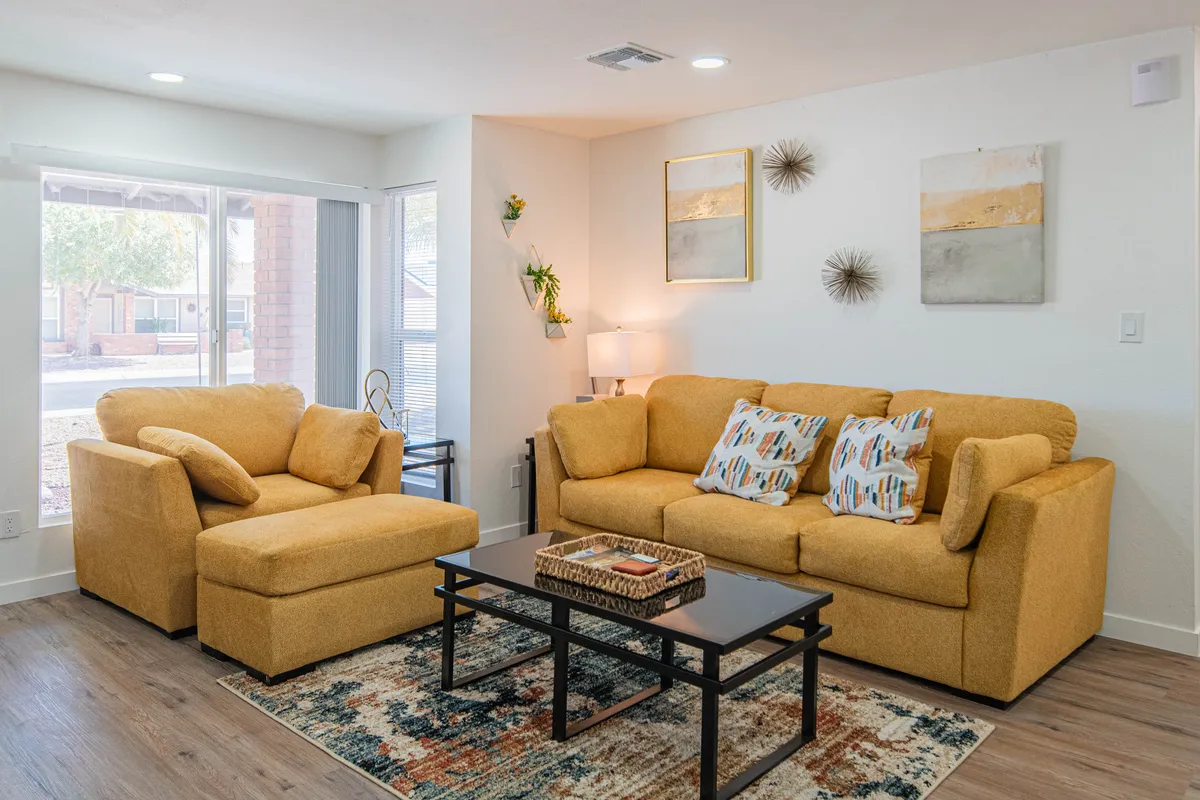 A cheerful living space with a yellow sofa, accent chair, coffee table, and striking wall art. The arrangement fosters both intimate conversations and group gatherings, with sunlight enhancing the cozy, vibrant mood.