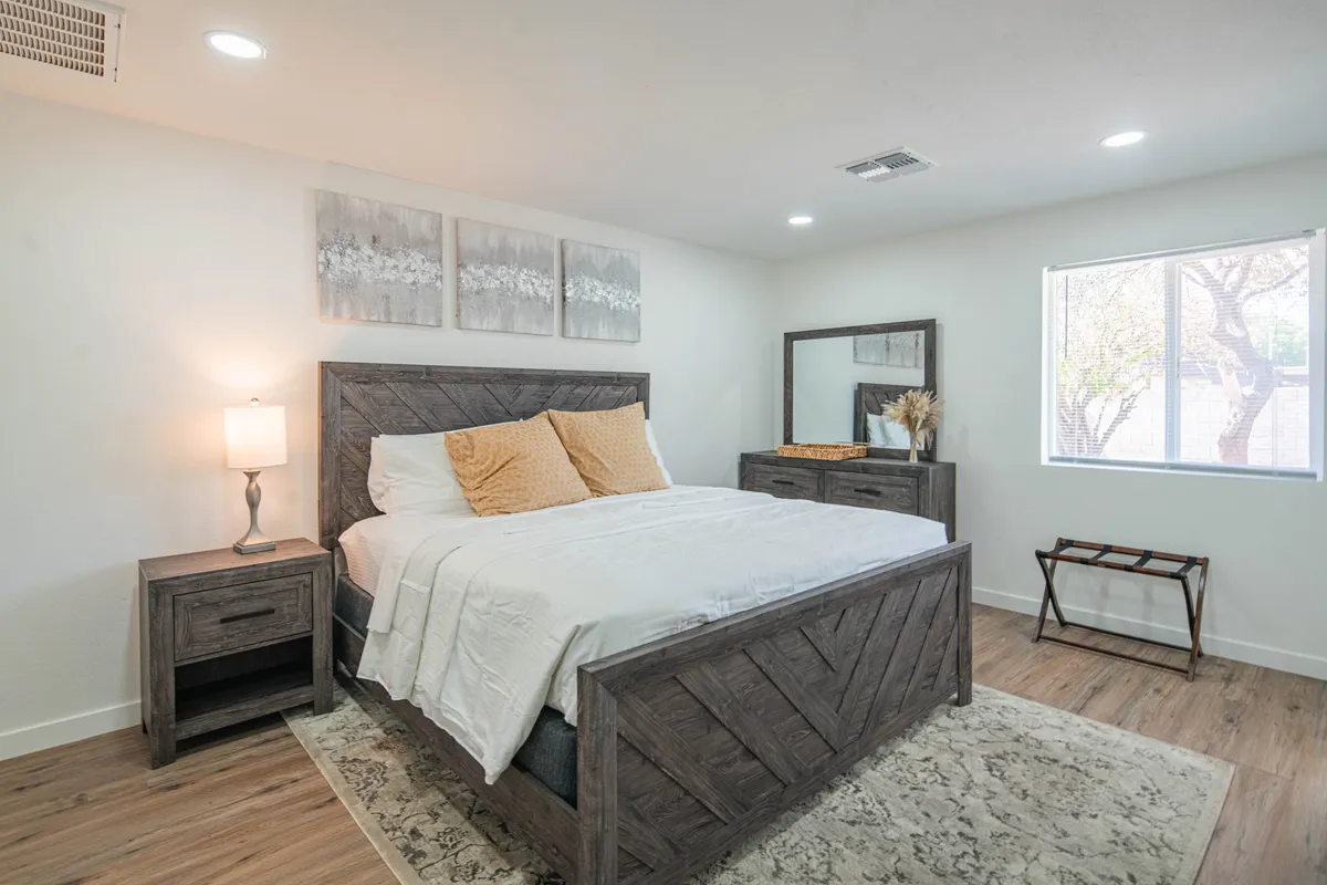 This image features a serene bedroom with a comfortable bed, nightstand, mirror, artwork, and a cozy rug. Natural light streams through the window, creating a calm and restful atmosphere perfect for relaxation or quiet activities.