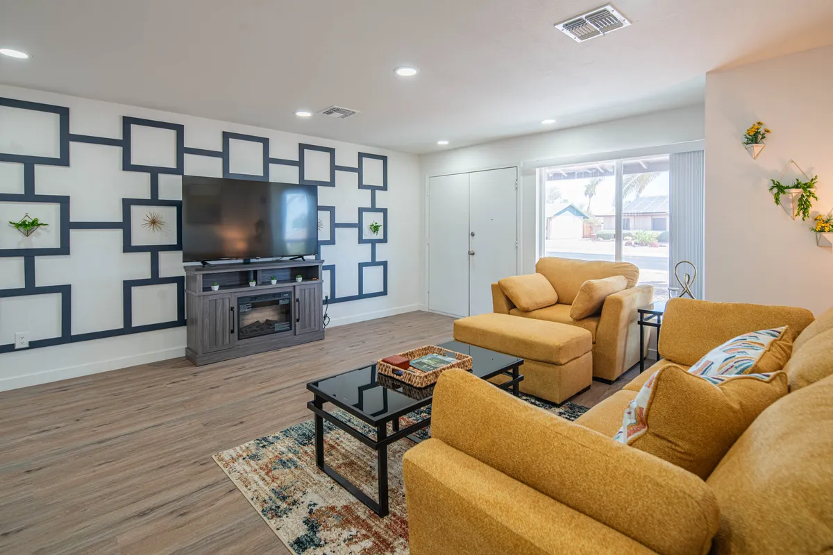 The image showcases a modern, inviting living space featuring a striking geometric-patterned accent wall, a large flat-screen TV, and plush, comfortable seating arranged for relaxation or gathering. The open design encourages conversation and connection, with ambient lighting creating a warm, cozy atmosphere—ideal for family movie nights or celebrating special occasions together.