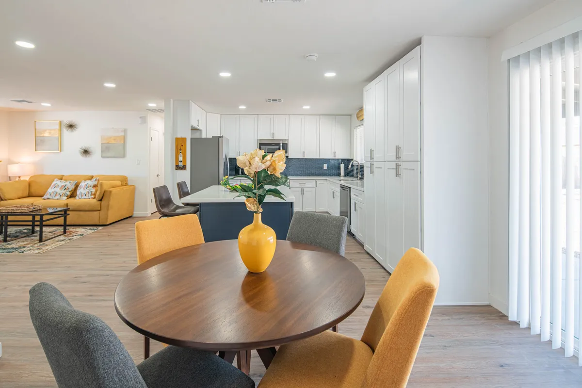 This image showcases a round dining table with a vase of flowers, a yellow couch, and the kitchen area in the background. The open layout offers a comfortable, inviting space suitable for working remotely or enjoying coffee breaks. Abundant natural light and an airy atmosphere suggest a pleasant workspace for productivity and relaxation.