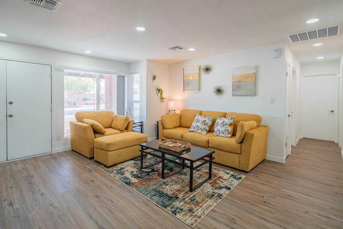 A bright and spacious living area featuring comfortable yellow couches, a colorful area rug, coffee table with decorative pieces, and artwork adorning the walls. Generous windows flood the room with natural light, and the open arrangement encourages movement and connection.