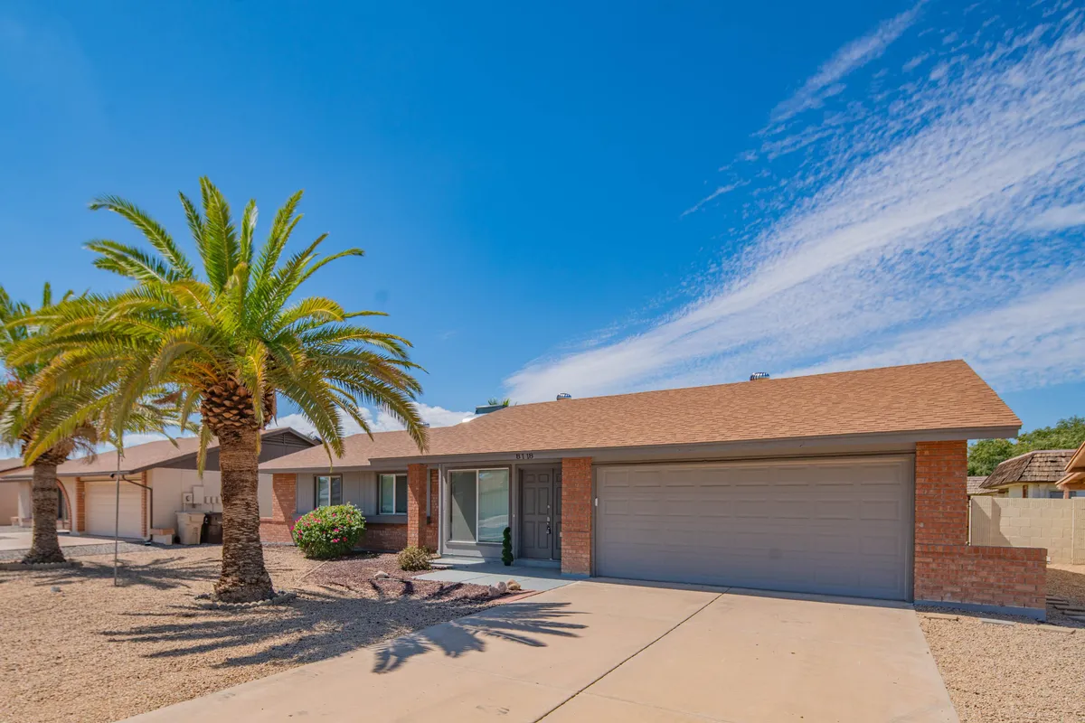 The exterior of Villa Theresa features a relaxed front yard with palm trees, a brick façade, and a garage door. The open space hints at the potential for outdoor gatherings or casual group activities under the Arizona sun.