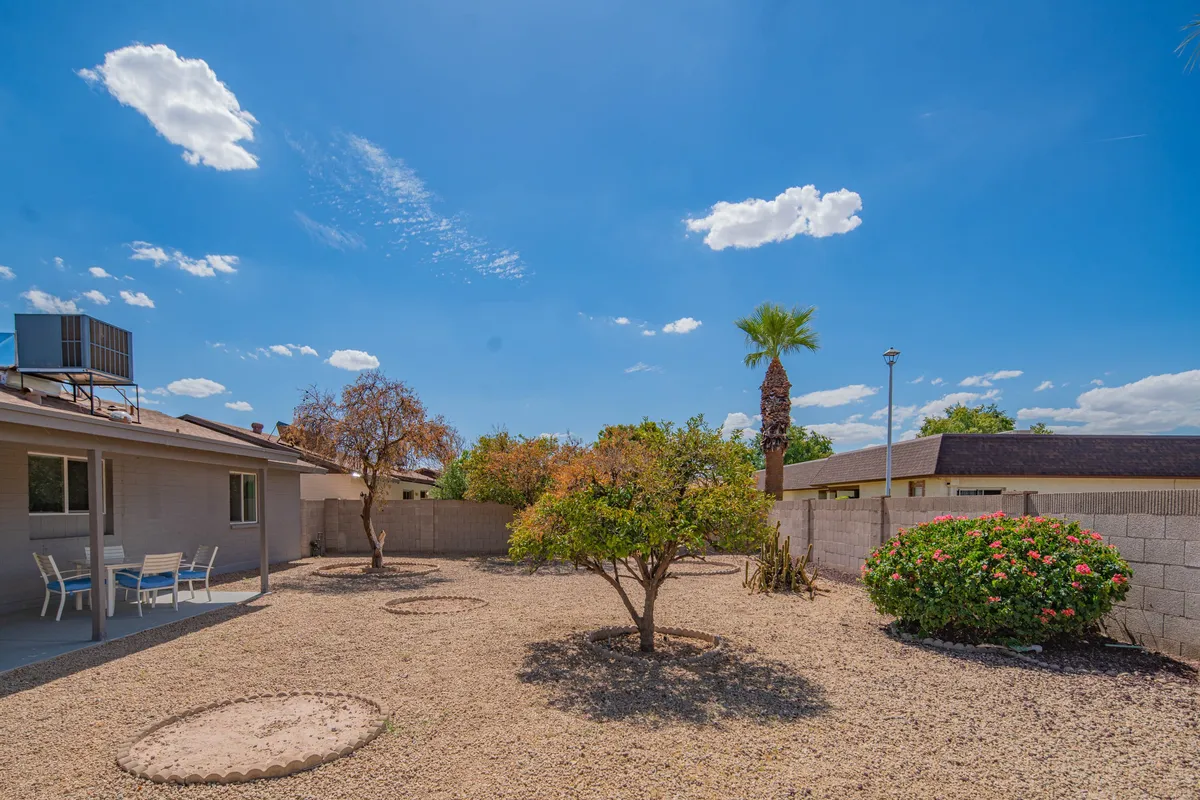 A tranquil garden scene with green trees, flowering bushes, gravel landscape, and a wide blue sky above. The space suggests relaxation and outdoor play, with enough room for children to explore and families to unwind together.