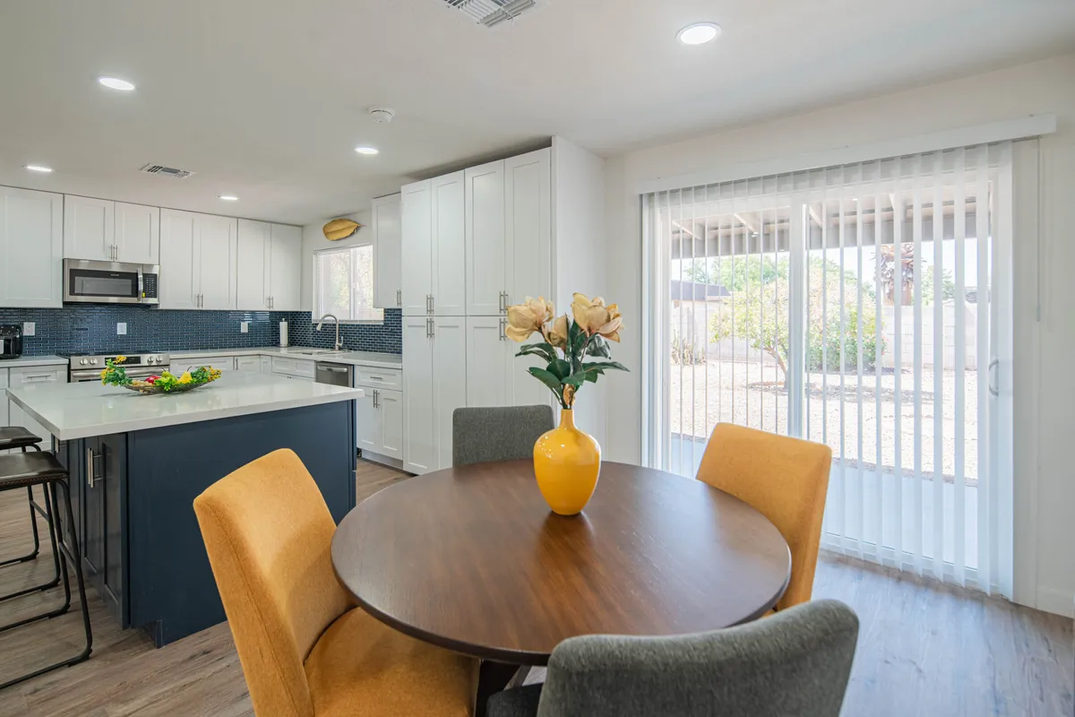 A warm, inviting dining space with a table set with flowers, a kitchen island nearby, and large windows flooding the room with natural light. The view encompasses both the dining and kitchen areas, showing how interconnected these spaces are.