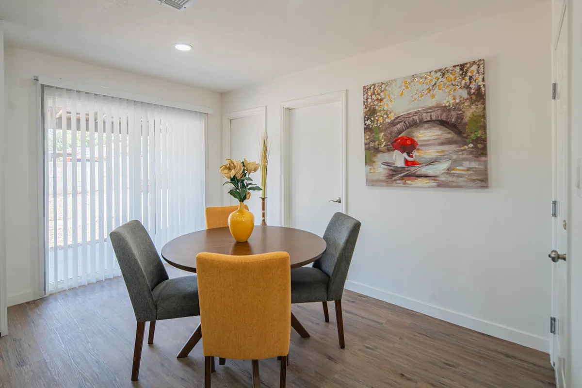 The photo features a cozy round dining table surrounded by colorful chairs, a cheerful floral centerpiece, and a painting of a bridge on the wall. The intimate, vibrant space encourages informal meetings, coffee chats, or quiet reflection during work breaks.